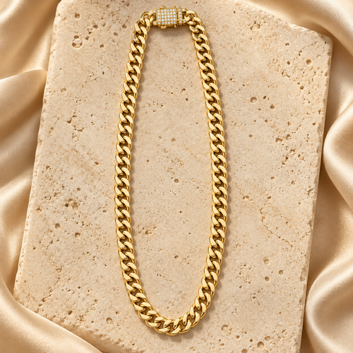 Cadena cubana de eslabones lisos en tono dorado con cierre rectangular decorado con piedras transparentes, sobre piedra crema y satén.