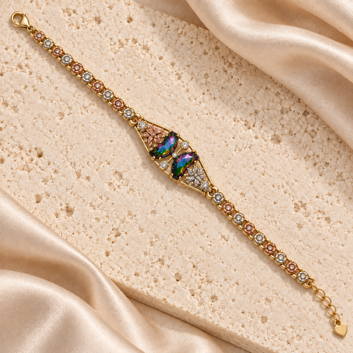 Pulsera dorada con dos piedras facetadas iridiscentes, detalles florales en tonos rosado y plateado, y extensión con dije de corazón.