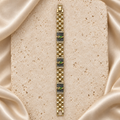 Una pulsera en tono dorado con tres placas de fondo negro, figuras de San Judas, filas de piedras transparentes y cierre plegable.