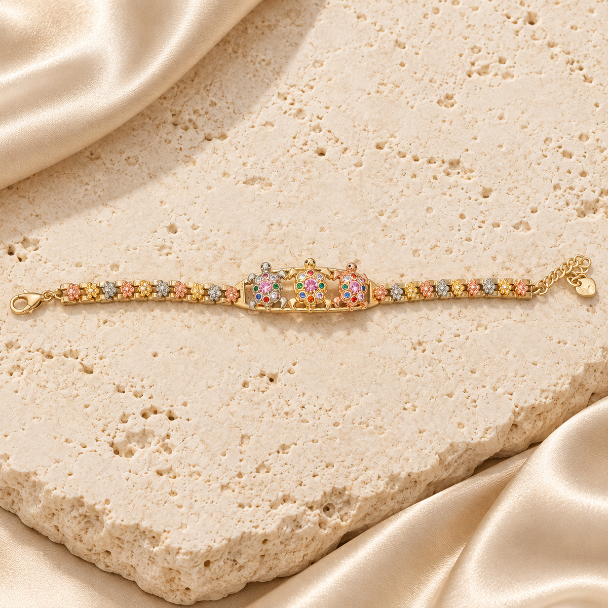 Una pulsera con tres tortugas de piedras multicolor, eslabones florales en tonos dorado, rosado y plateado, cierre de langosta y extensión con corazón.