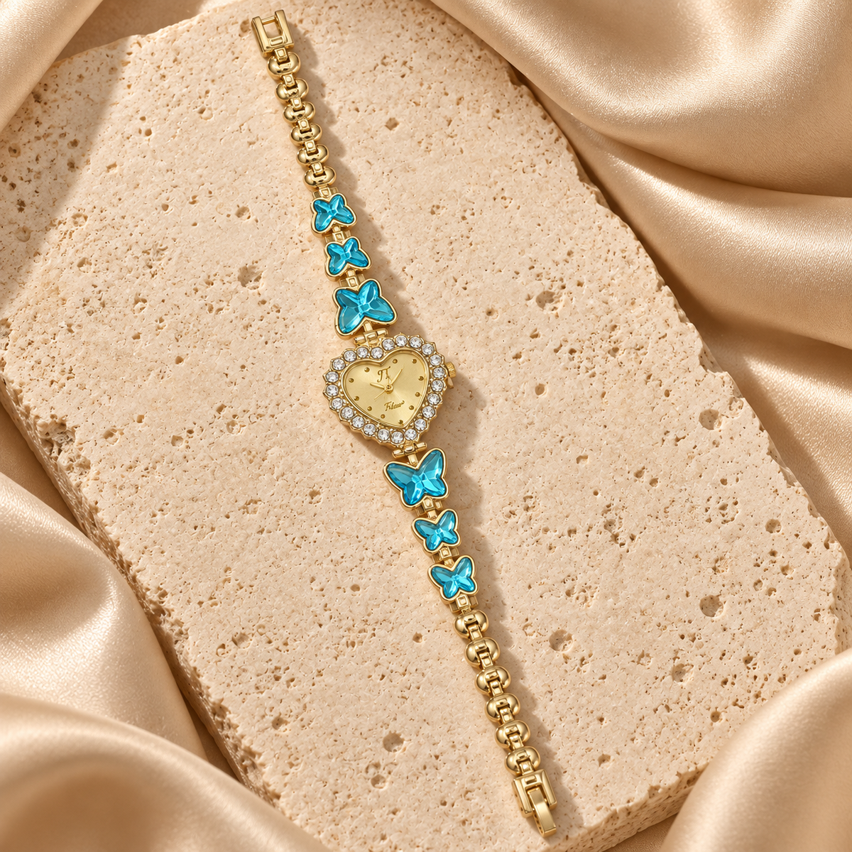 Reloj de pulsera dorado con carátula de corazón rodeada de piedras transparentes y seis mariposas turquesas.