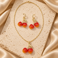 Collar de cadena dorada con dije de dos cerezas rojas y naranjas, junto a aretes de cerezas con pequeños aros brillantes.