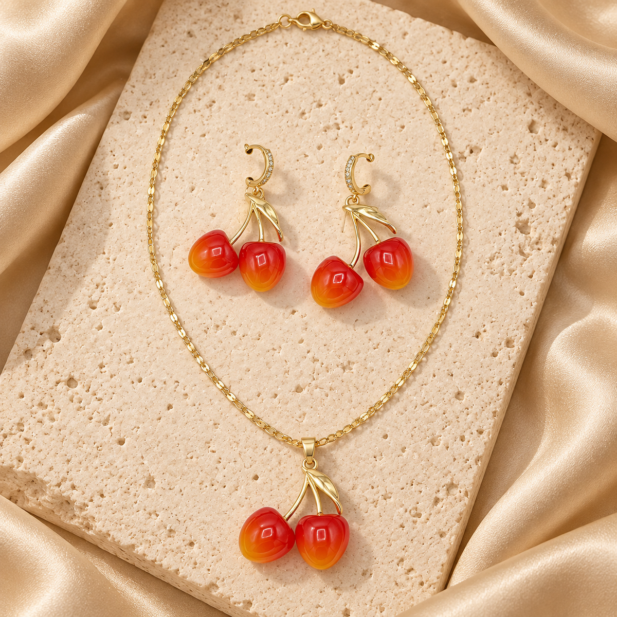Collar de cadena dorada con dije de dos cerezas rojas y naranjas, junto a aretes de cerezas con pequeños aros brillantes.