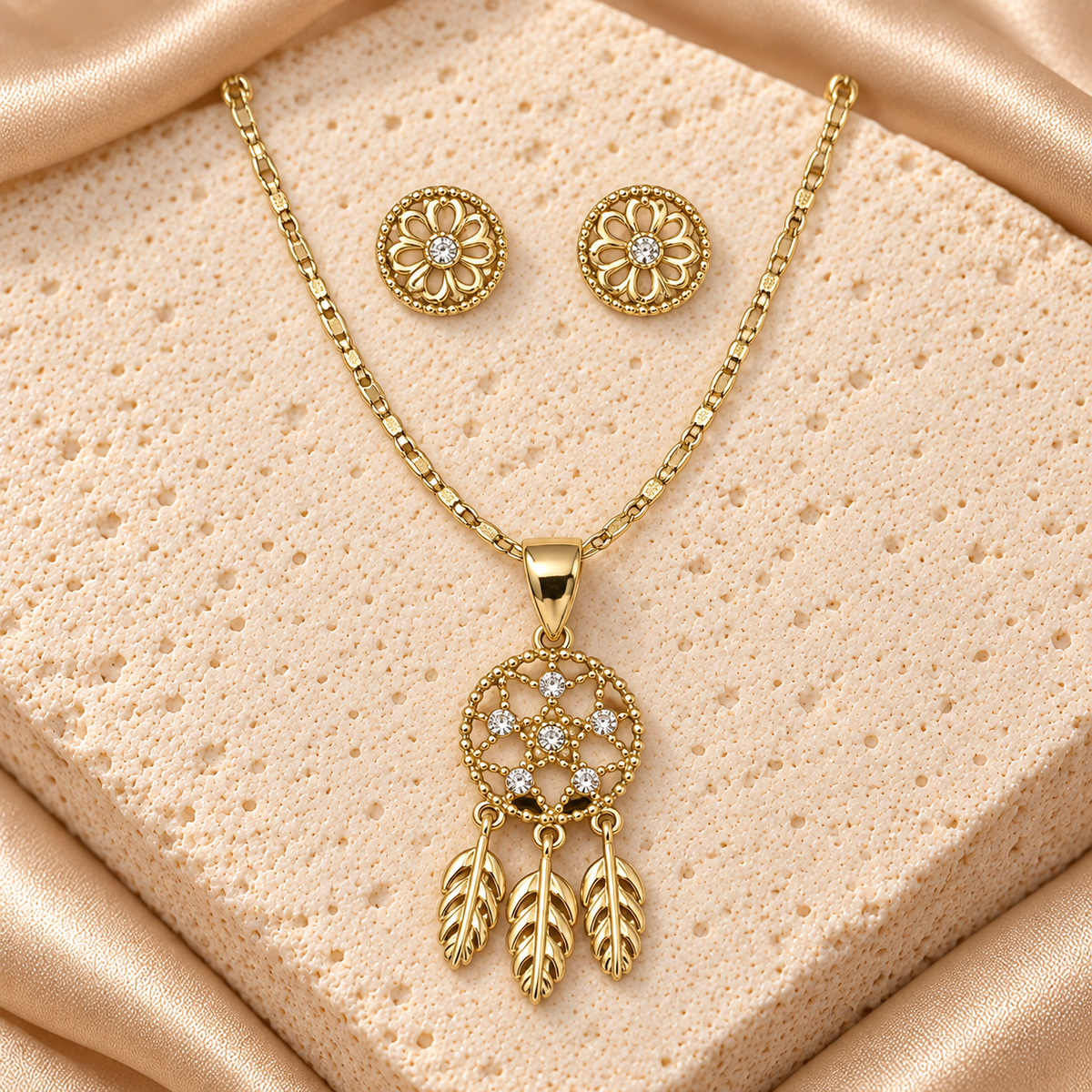 Collar en tono dorado con dije circular calado, seis piedras transparentes y tres plumas anchas colgantes, junto a dos aretes florales redondos.