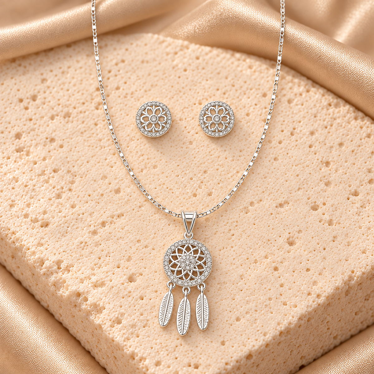 Collar en tono plateado con dije circular de atrapasueños, tres plumas colgantes y un par de aretes redondos florales con detalles brillantes.