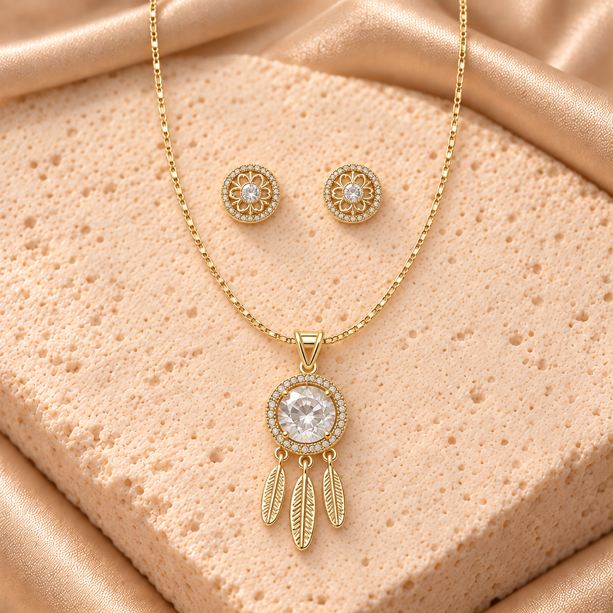 Set en tono dorado: collar con piedra redonda transparente, halo brillante y tres plumas colgantes, acompañado de dos aretes florales redondos.