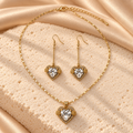 Set dorado de collar y dos aretes largos de gancho con corazones transparentes, bordes brillantes y detalles laterales texturizados.